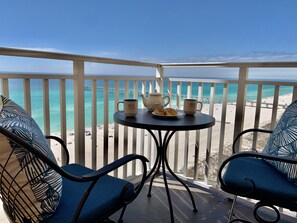Ein Balkon mit Blick auf den Strand, ein Tisch mit Kaffeetassen und einem Teekessel sowie zwei Stühle mit tropischem Muster.