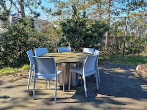 A round wooden table with six white chairs, surrounded by dense greenery and trees. - Vacation home for 6 people located directly on the dune (Wangerooge)