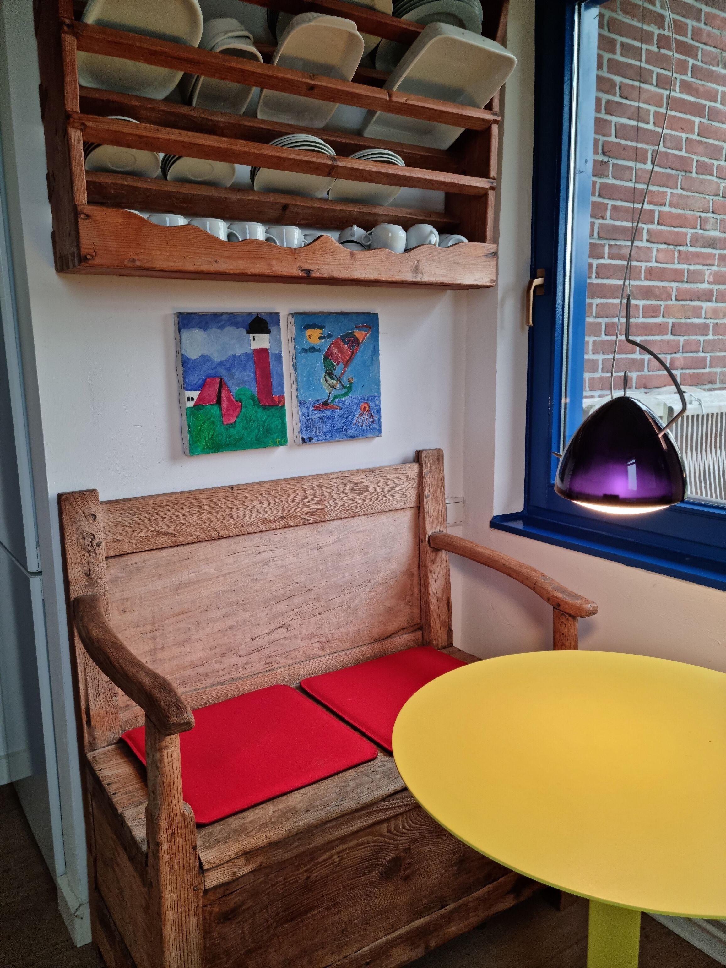 A wooden bench with red cushions, a yellow table, and a shelf with dishes and plates.