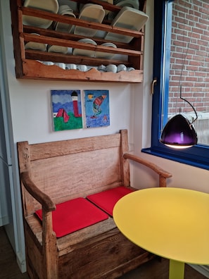 A wooden bench with red cushions, a yellow table, and a shelf with dishes and plates. - Vacation home for 6 people located directly on the dune (Wangerooge)