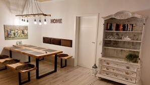 En matplats med ett träbord och bänkar, en väggmonterad lampa, en bokhylla med böcker och dekorativa föremål samt en dörr.