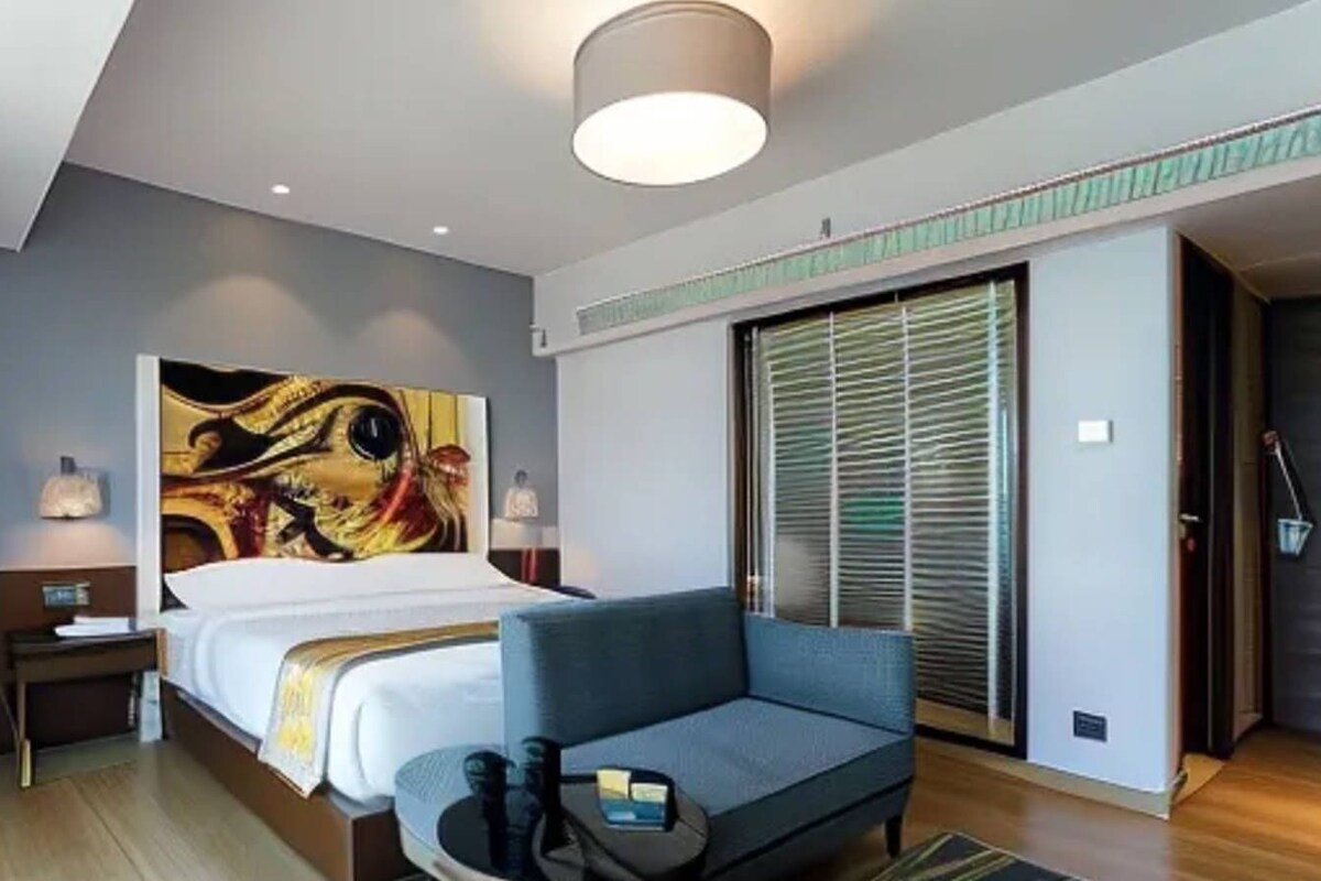 Una habitación de hotel moderna con una cama grande, un sofá azul, una obra de arte en la pared, una mesita de noche con lámpara y un armario con puerta corredera.