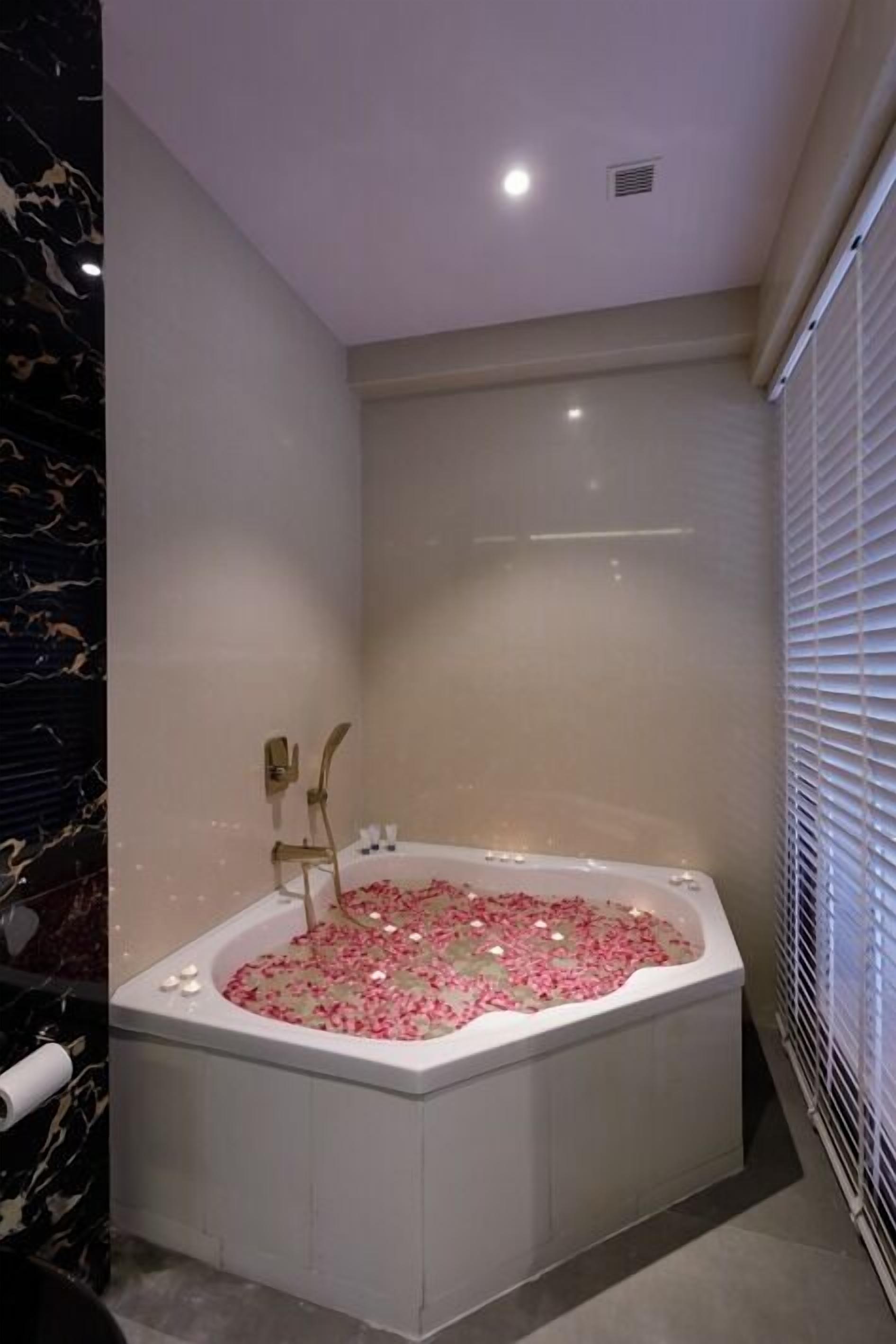 A modern bathroom with a freestanding bathtub filled with rose petals, a marble wall, and a window with blinds.