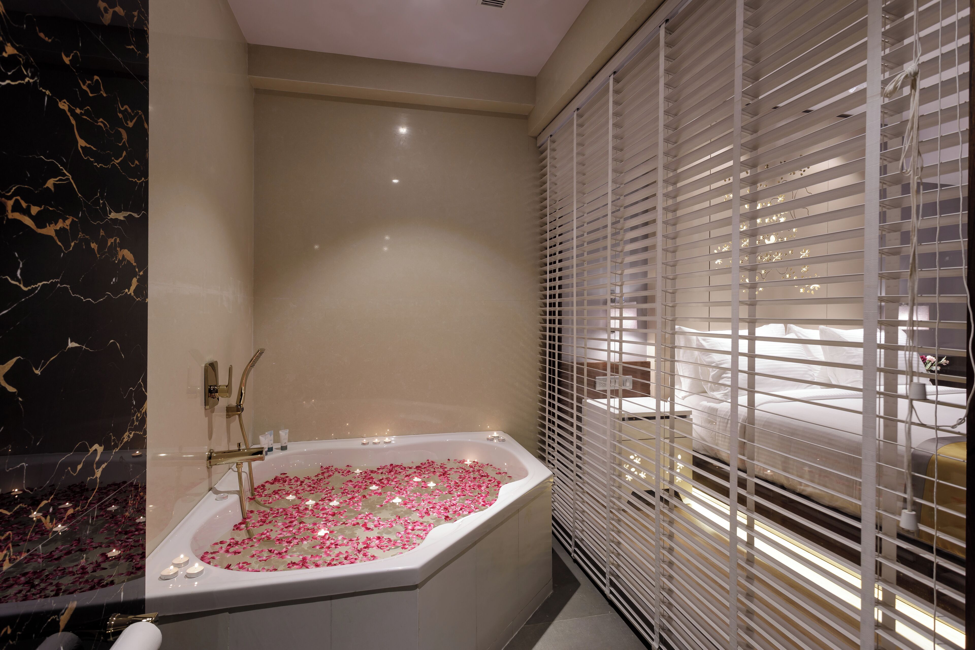 A modern bathroom with a freestanding bathtub filled with rose petals, a marble wall, and a window with blinds.