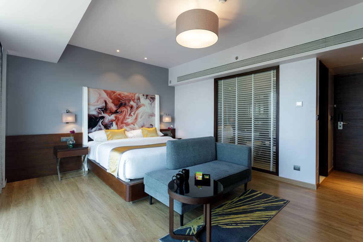 Quarto de hotel moderno com uma cama, um sofá, uma mesinha e um grande quadro abstrato na parede.