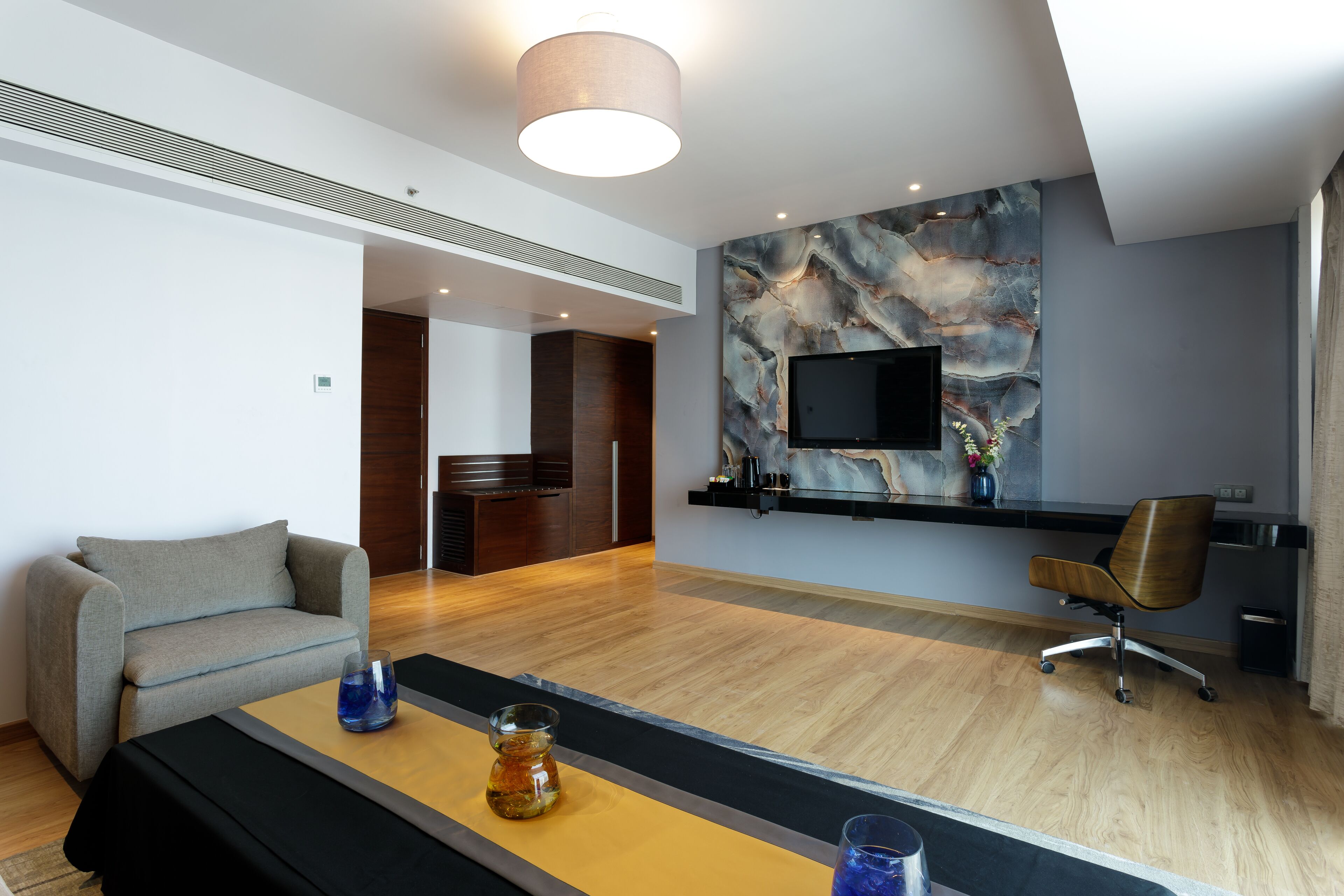 A modern hotel room with a wooden floor, a grey sofa, a black coffee table, and a TV mounted on the wall.