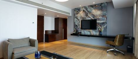 A modern hotel room with a wooden floor, a grey sofa, a black coffee table, and a TV mounted on the wall.