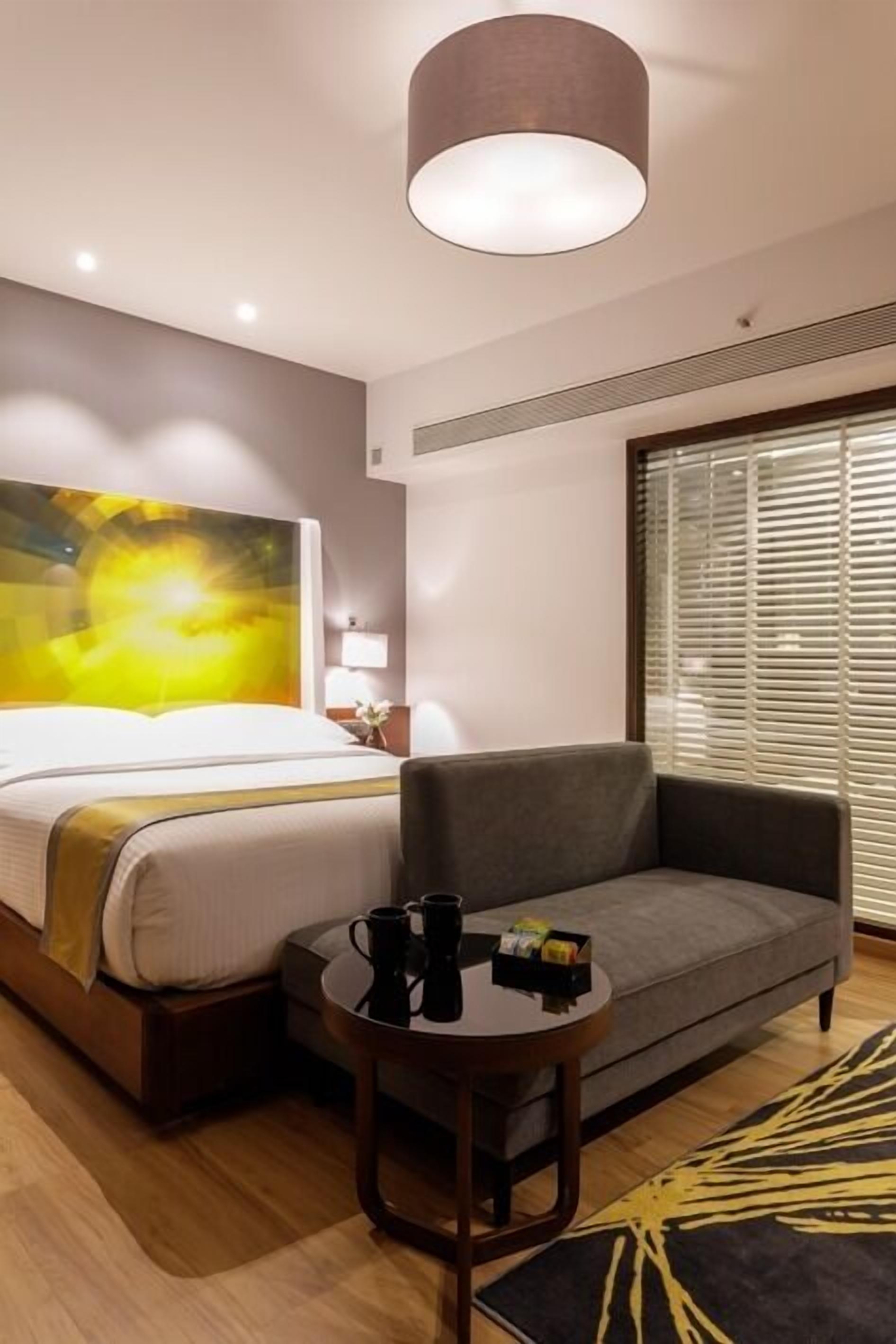 A hotel room with a bed, a gray sofa, a small table with a coffee cup, and a large window with blinds.