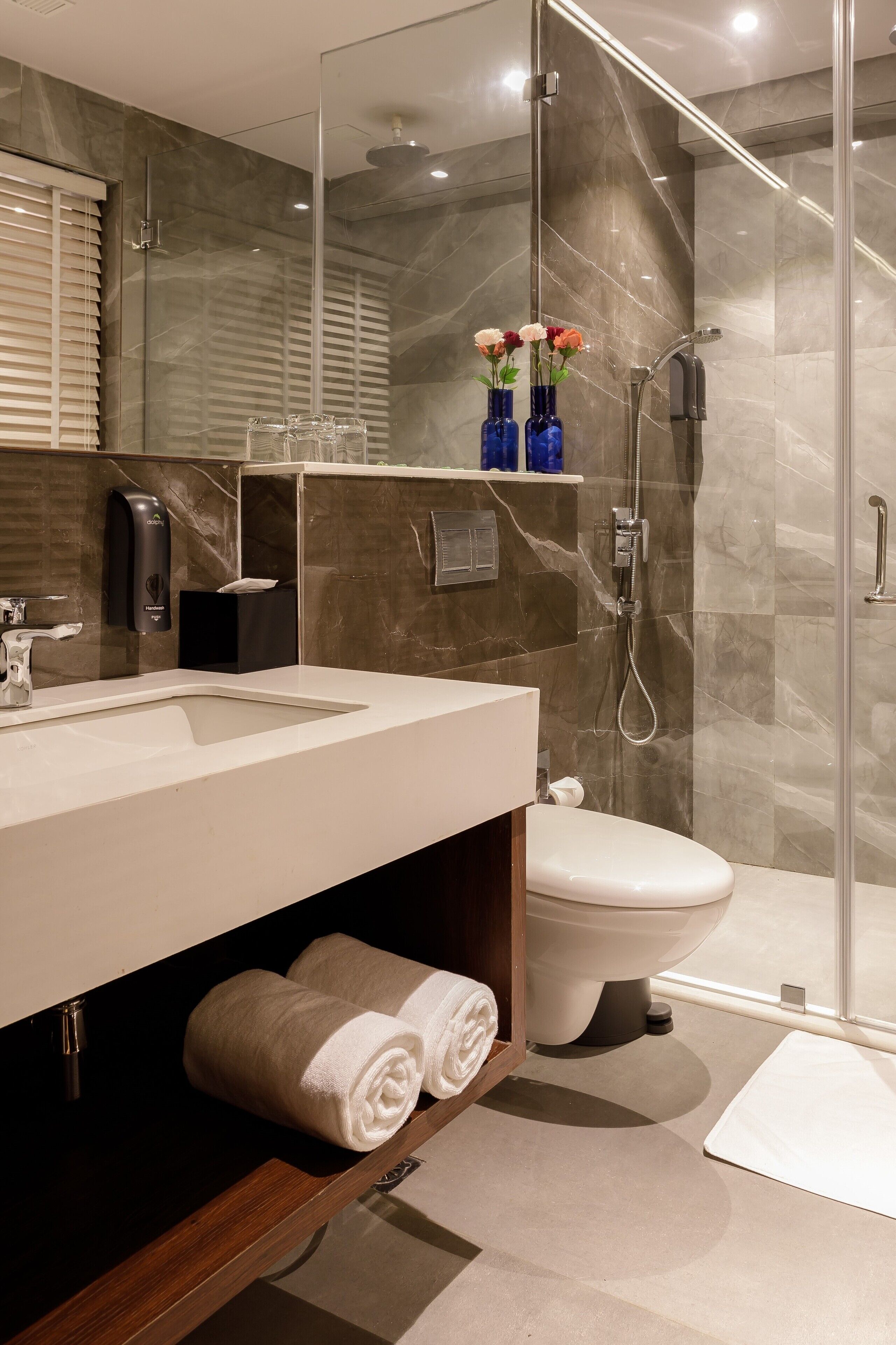 A modern bathroom with a glass-enclosed shower, a white sink, and a toilet. There are towels on a shelf and a vase with flowers on the counter.