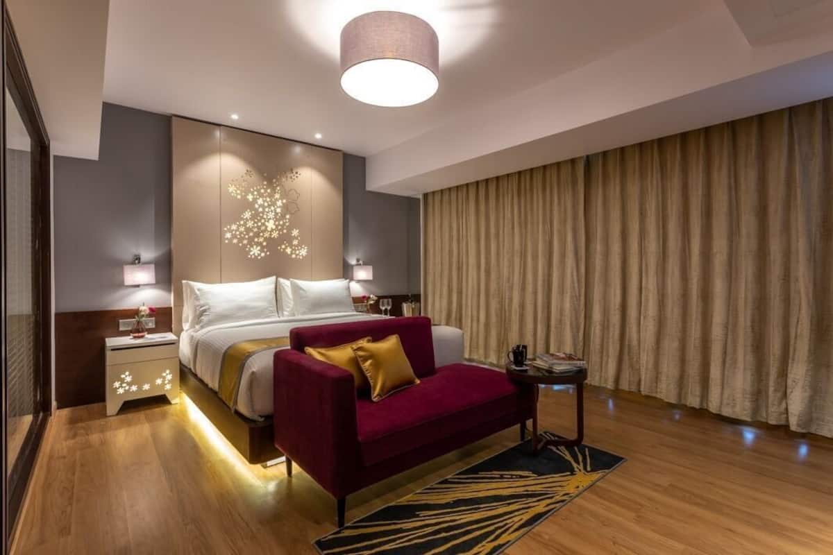 Quarto de hotel moderno com uma cama grande, um sofá vermelho, uma mesinha de apoio e uma janela ampla com cortinas.