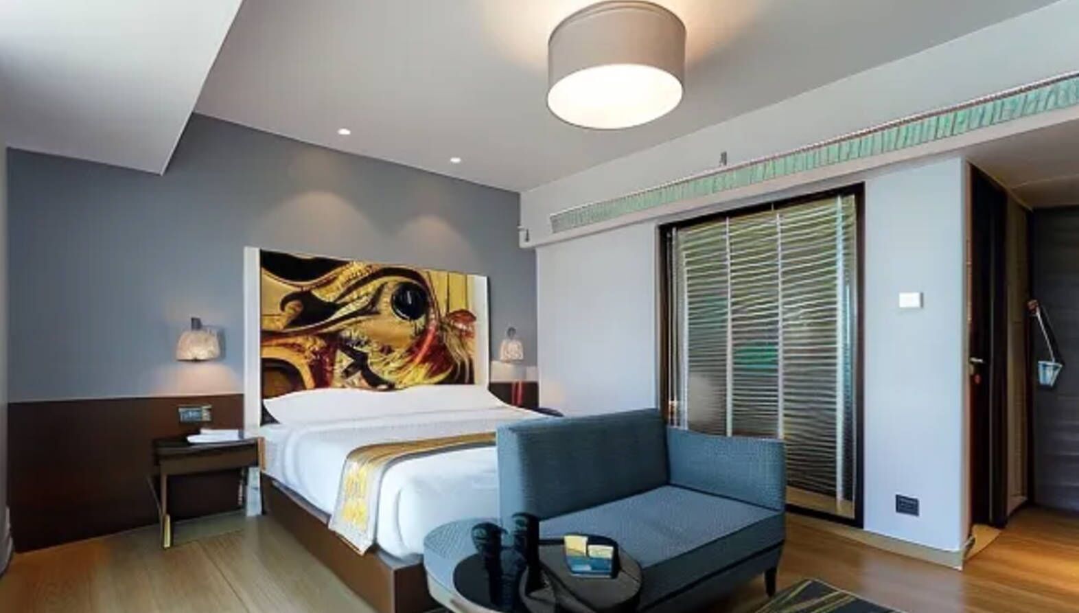 A modern hotel room with a large bed, a blue sofa, a wall-mounted artwork, a bedside table with a lamp, and a sliding door closet.