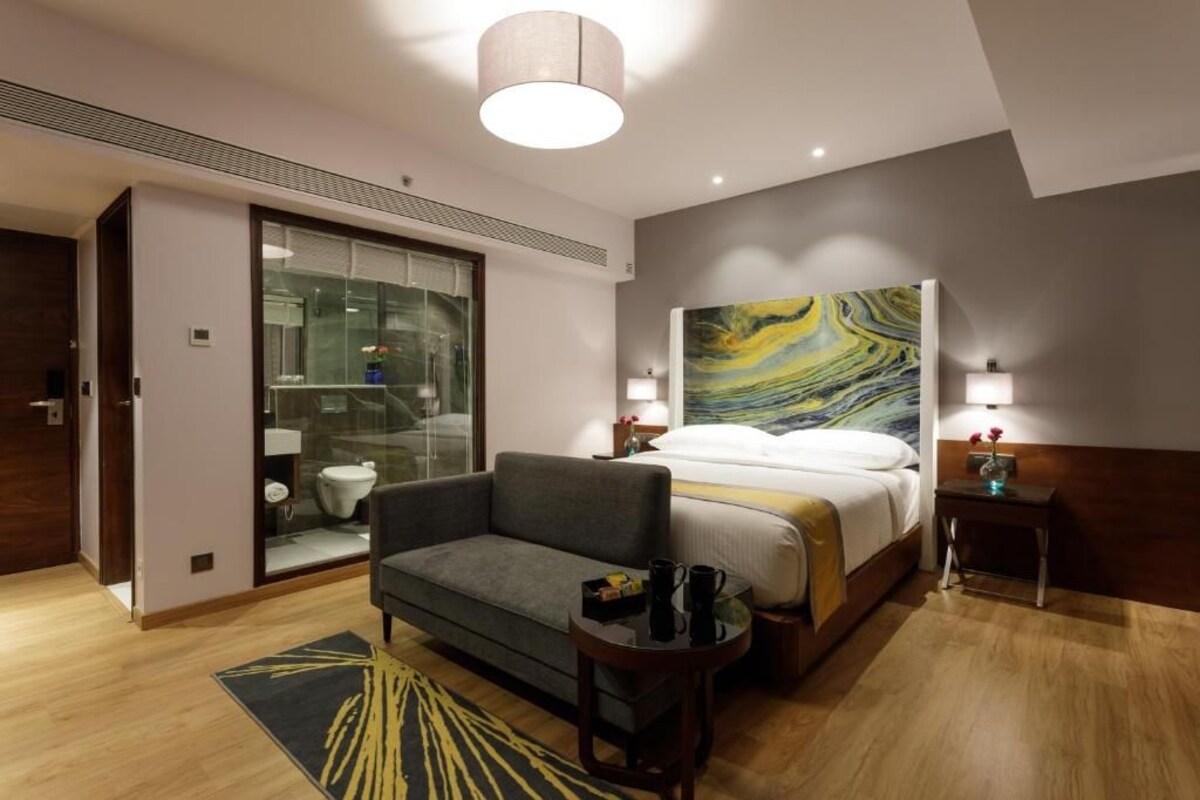 Una habitación de hotel moderna con una cama, un sofá, una mesita y un baño con paredes de vidrio.