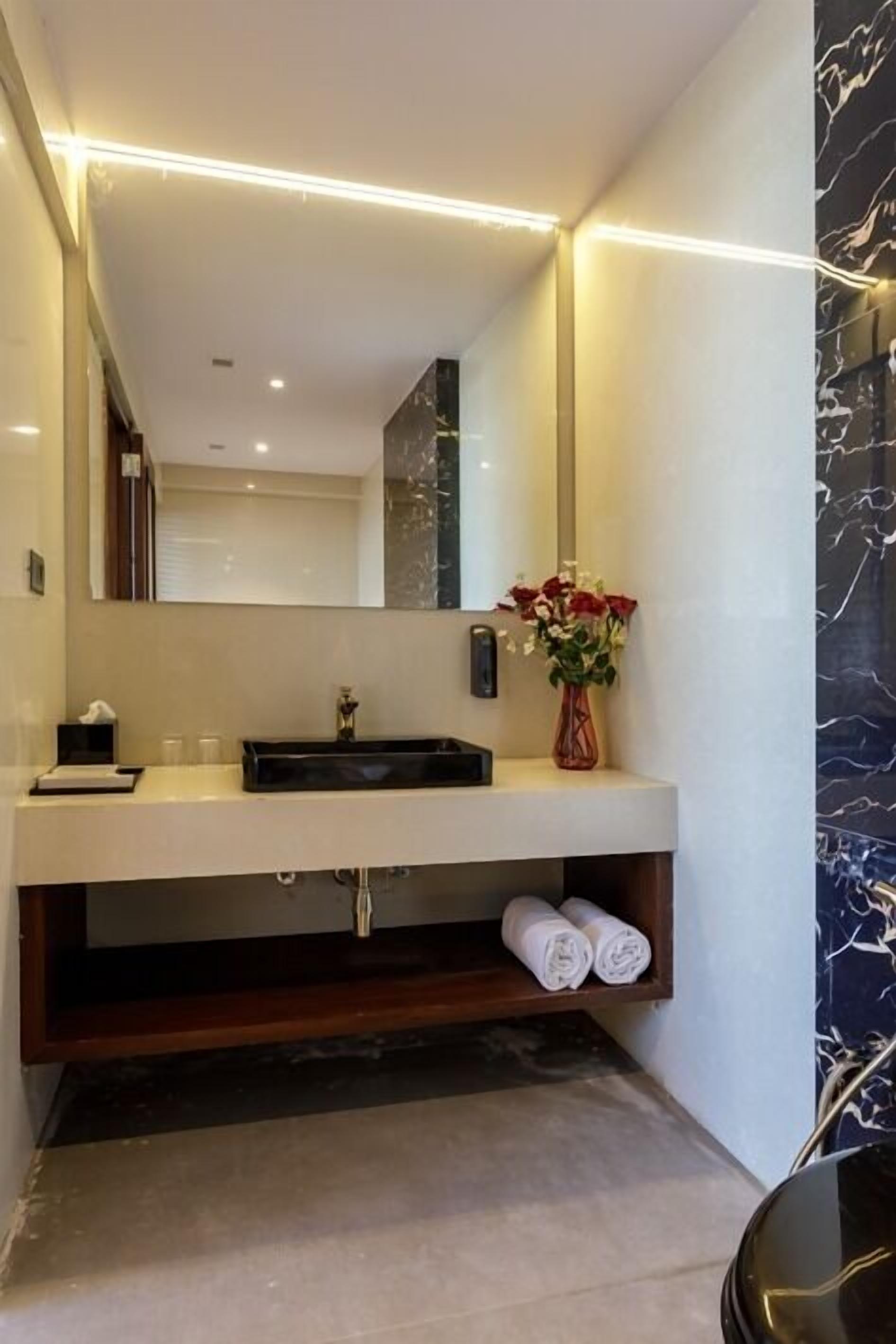 Un baño moderno con un tocador blanco, un lavabo negro y un salpicadero de mármol. Hay un espejo sobre el lavabo y un jarrón con flores en el tocador.