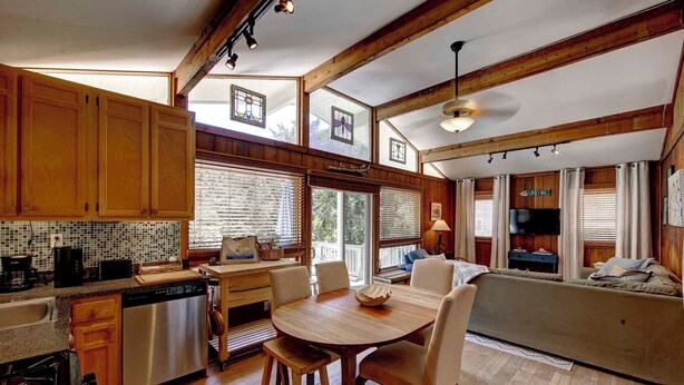 A spacious kitchen with wooden cabinets, a dining table with chairs, a built-in oven, and a large window with blinds. - Sandy Pause - Ocracoke Island (Ocracoke)