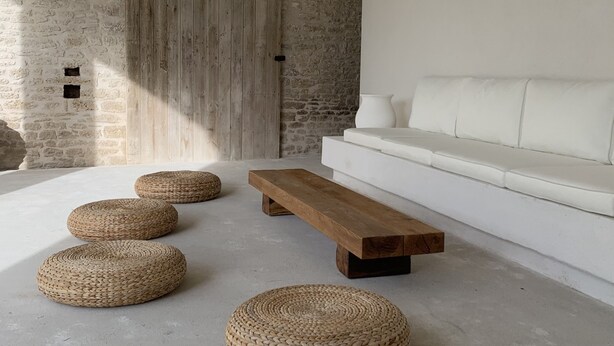 Un salon moderne avec un canapé blanc, un banc en bois et des poufs en fibres naturelles, le tout posé sur un sol en béton.