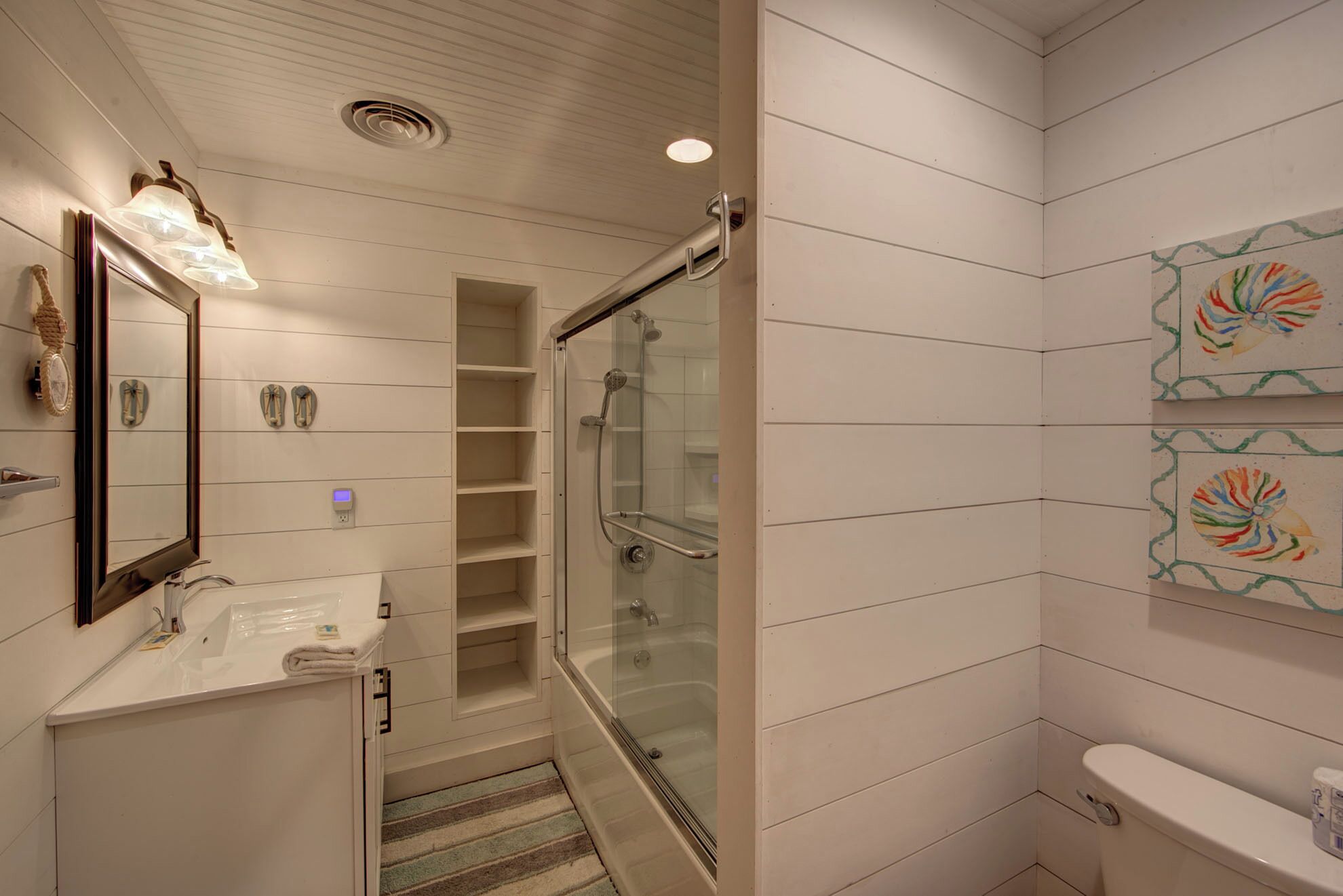 A bathroom with a glass-enclosed shower, a white sink, and a toilet. There are shelves and a mirror on the wall.