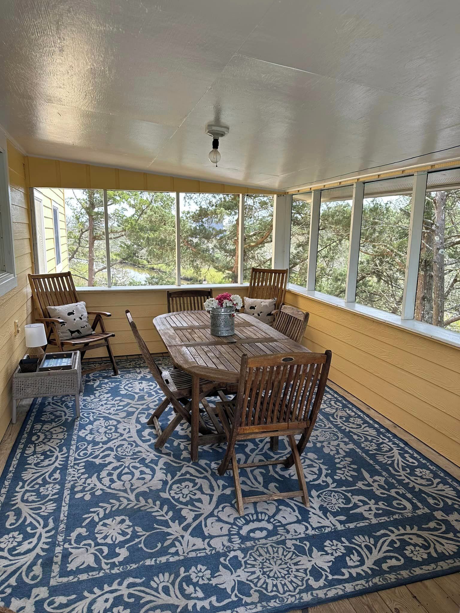 Une véranda avec une table et des chaises en bois, de grandes fenêtres et un tapis à motifs bleus.