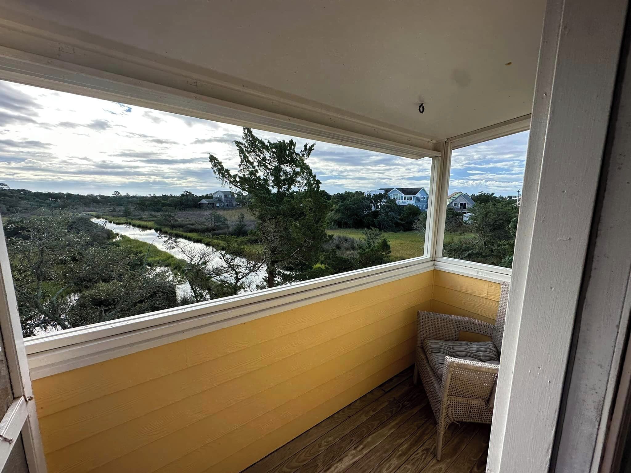 A balcony with a view of a river and houses, wooden flooring, and a wicker chair.