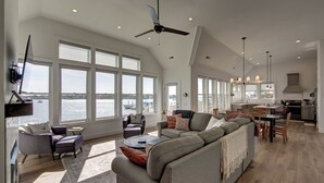 A spacious living area with a large window overlooking a body of water, a ceiling fan, and a modern kitchen in the background.
