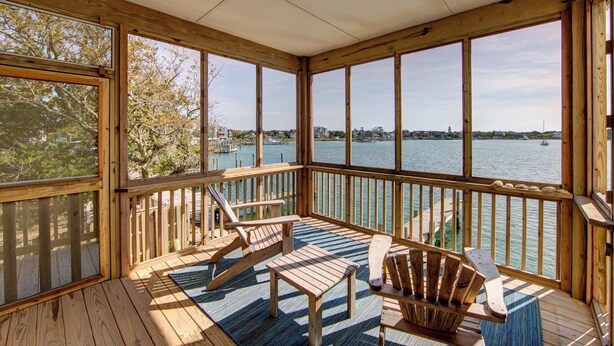 A wooden deck with a view of a body of water, featuring two Adirondack chairs and a small table.
