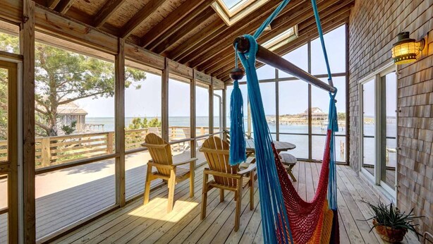 Una terraza de madera con una hamaca, sillas y una mesa, con vista a un cuerpo de agua.