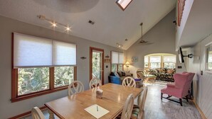 A spacious dining area with a wooden table and chairs, a cozy seating area with a sofa and chairs, and large windows offering views of the outdoors. - Charlotte's Place - Ocracoke Island (Ocracoke)