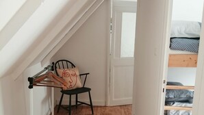 Pieni, kapea huone, jossa on kalteva katto, puulattia, metallinen pyykiteline, tuoli ja kuvioitu viltti päällä oleva sänky.