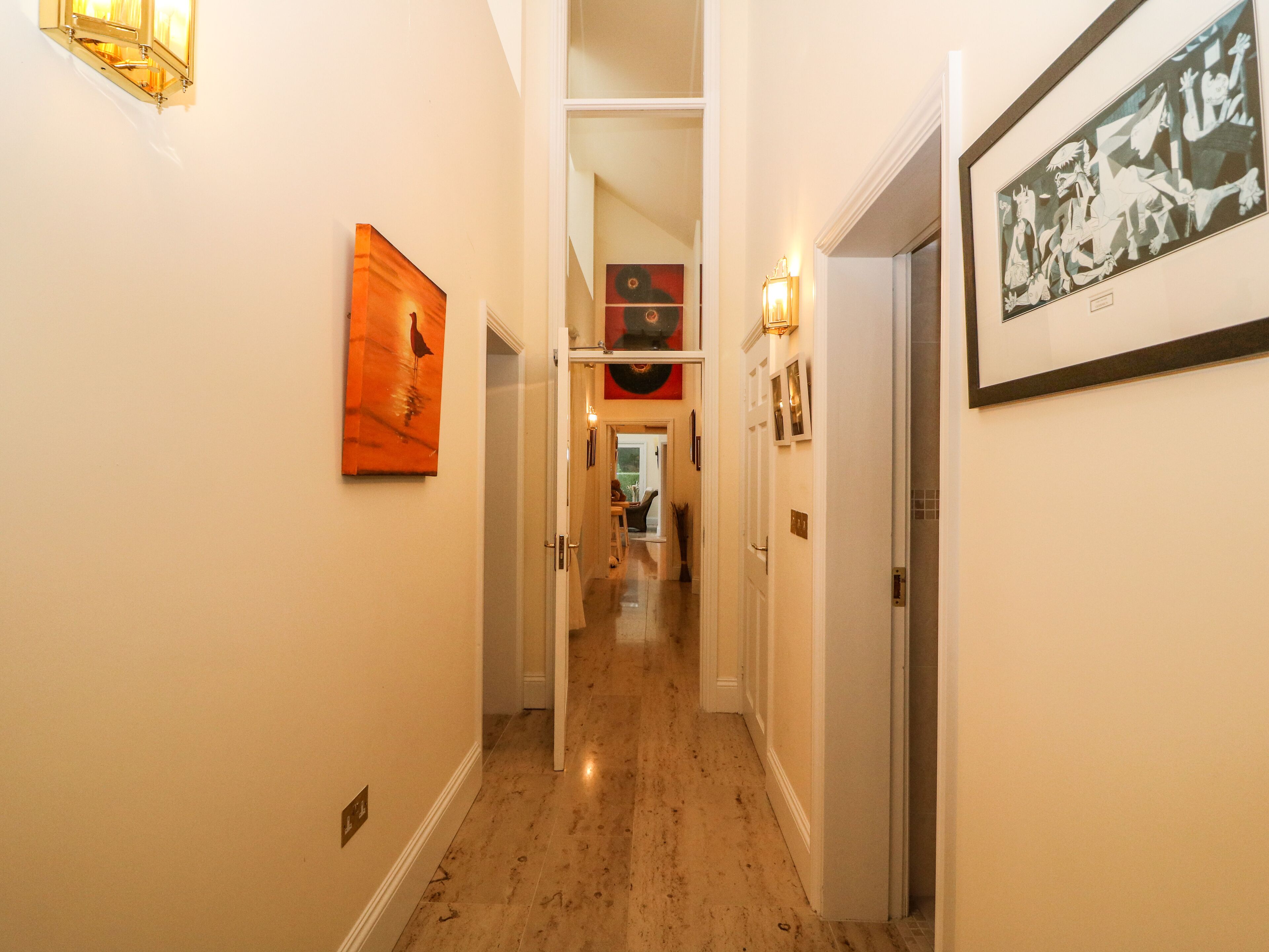 A hallway with artwork on the walls, a door on the right, and a view into another room.