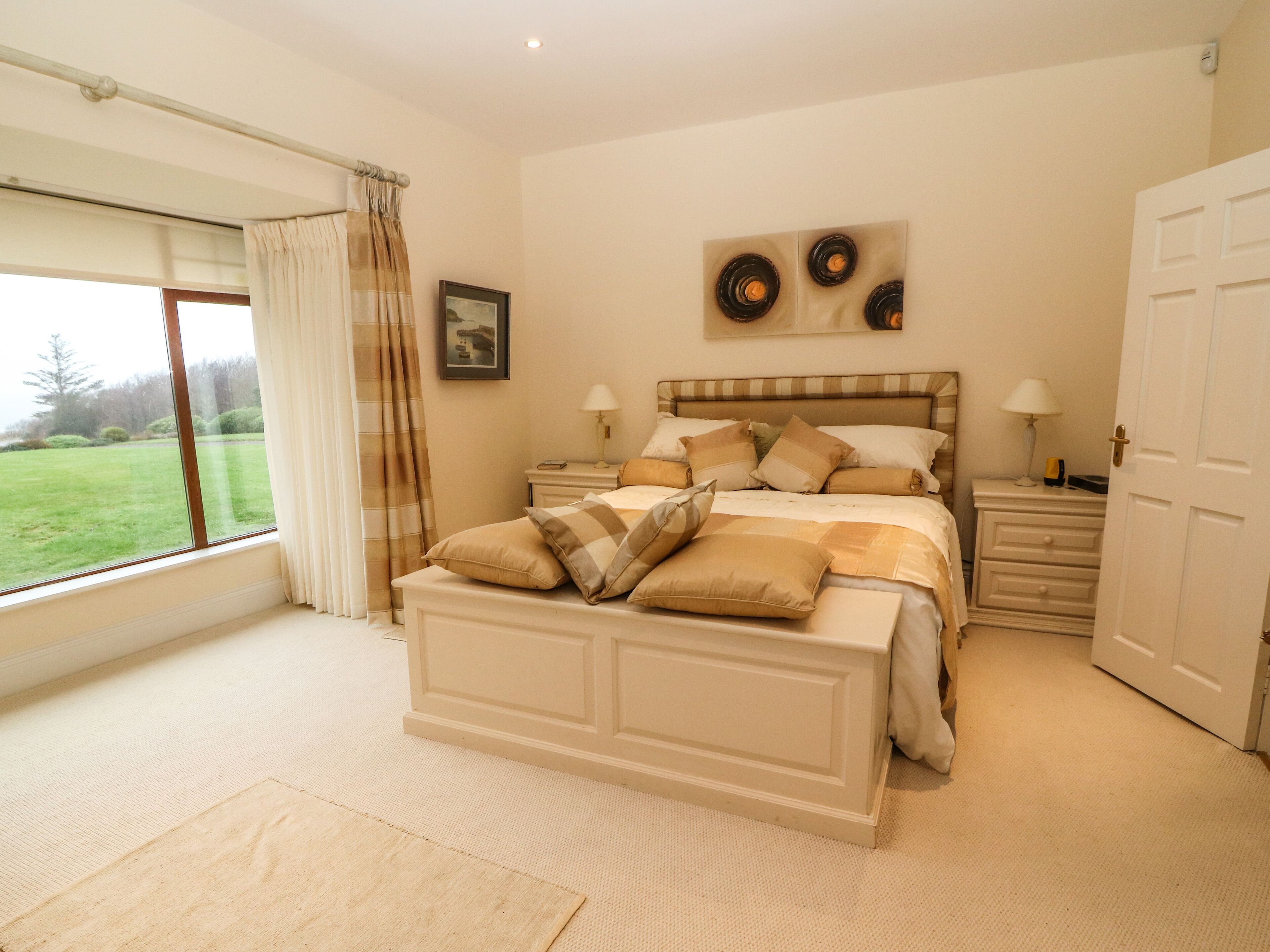A bedroom with a large bed, two bedside tables, a window with curtains, a door, and a wall-mounted artwork.