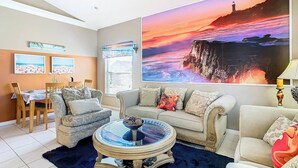 Un salon agrémenté d’un grand tableau représentant un coucher de soleil, un coin repas avec une table et des chaises, et un espace salon confortable avec un canapé et une table basse.
