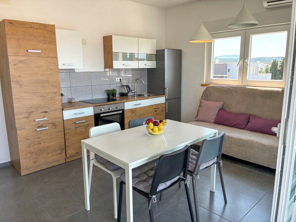 A compact kitchen with modern appliances, a dining area with a table and chairs, and a sofa with a view of the outdoors.