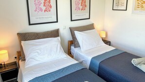 Ett hotellrum med två sängar, var och en med en kudde och ett täcke, ett sängbord med en lampa och inramade konstverk på väggen.