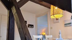 Un coin repas avec une table blanche, des chaises et une lampe jaune. On aperçoit une structure en poutres de bois au premier plan.