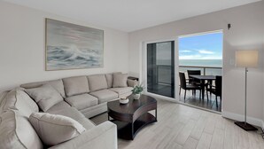 A modern living room with a large window, a sofa, a coffee table, and a view of the ocean. - BEACHFRONT ON A BUDGET!  MILLION $ VIEW!  2B/2B ISLAND VACATION CONDO! (Fort Myers Beach)