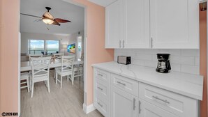 A modern kitchen with white cabinets, a ceiling fan, a dining area with white chairs, and a view to the outside. - BEACHFRONT ON A BUDGET!  MILLION $ VIEW!  2B/2B ISLAND VACATION CONDO! (Fort Myers Beach)