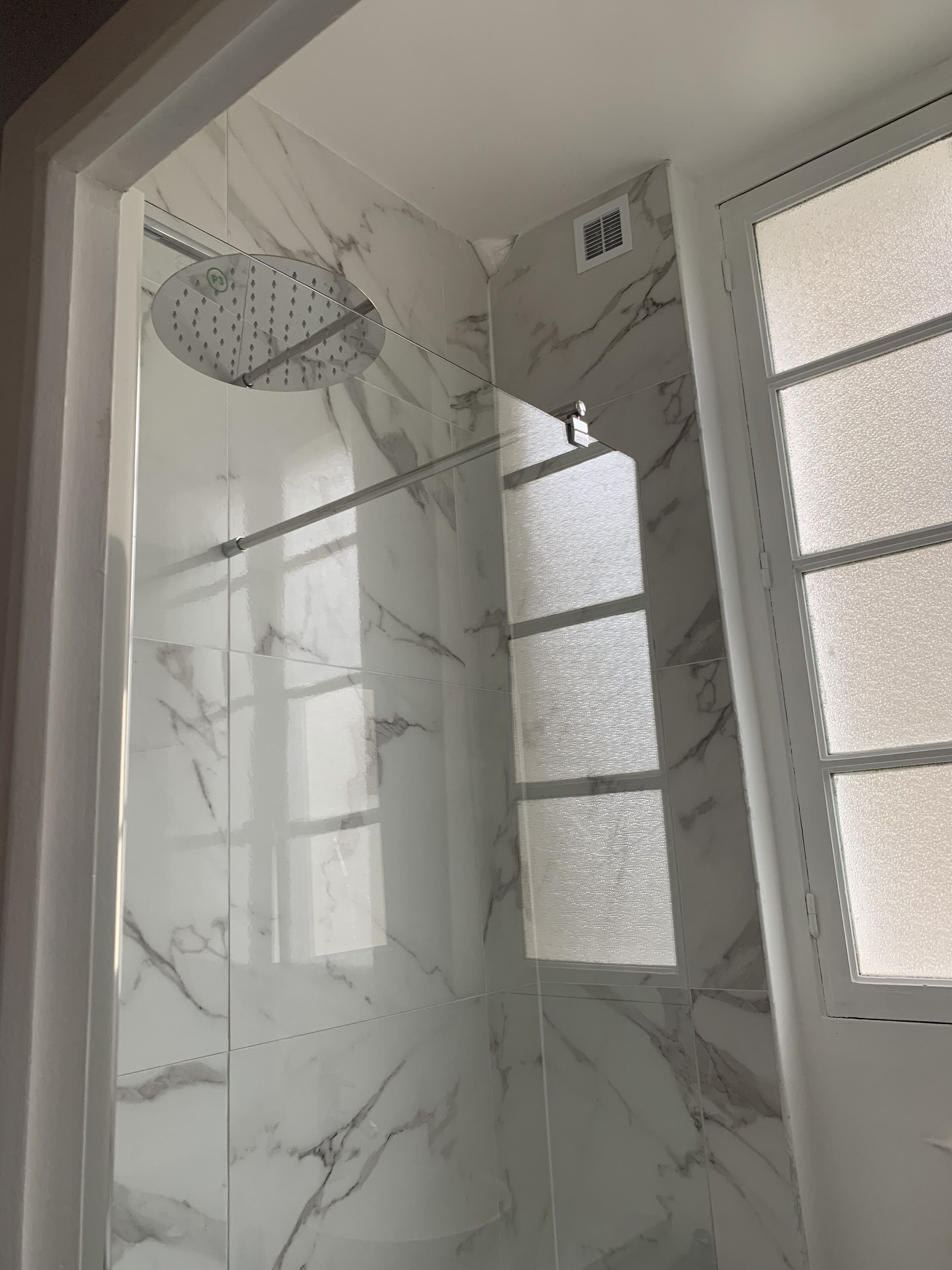 A bathroom with marble tiles, a shower with a rain showerhead, and frosted glass windows.