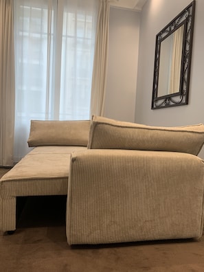 Un sofá color beige con un reposapiés a juego en una habitación con un ventanal grande, cortinas translúcidas y un espejo decorativo en la pared.