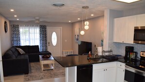 A modern kitchen with a black countertop, white cabinets, and stainless steel appliances. There is a black sofa and a small table in the living area.