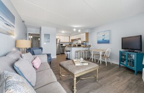 Una sala de estar con sofá, reposapiés y televisor. Se observa una cocina al fondo con zona de comedor.