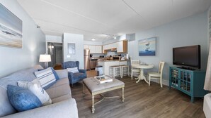 Un salon comprenant un canapé, un pouf et un téléviseur. Il y a également un coin cuisine avec un réfrigérateur, un four à micro-ondes et une table à manger.