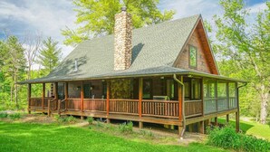 A log cabin with a stone chimney, wooden deck, and a covered porch.