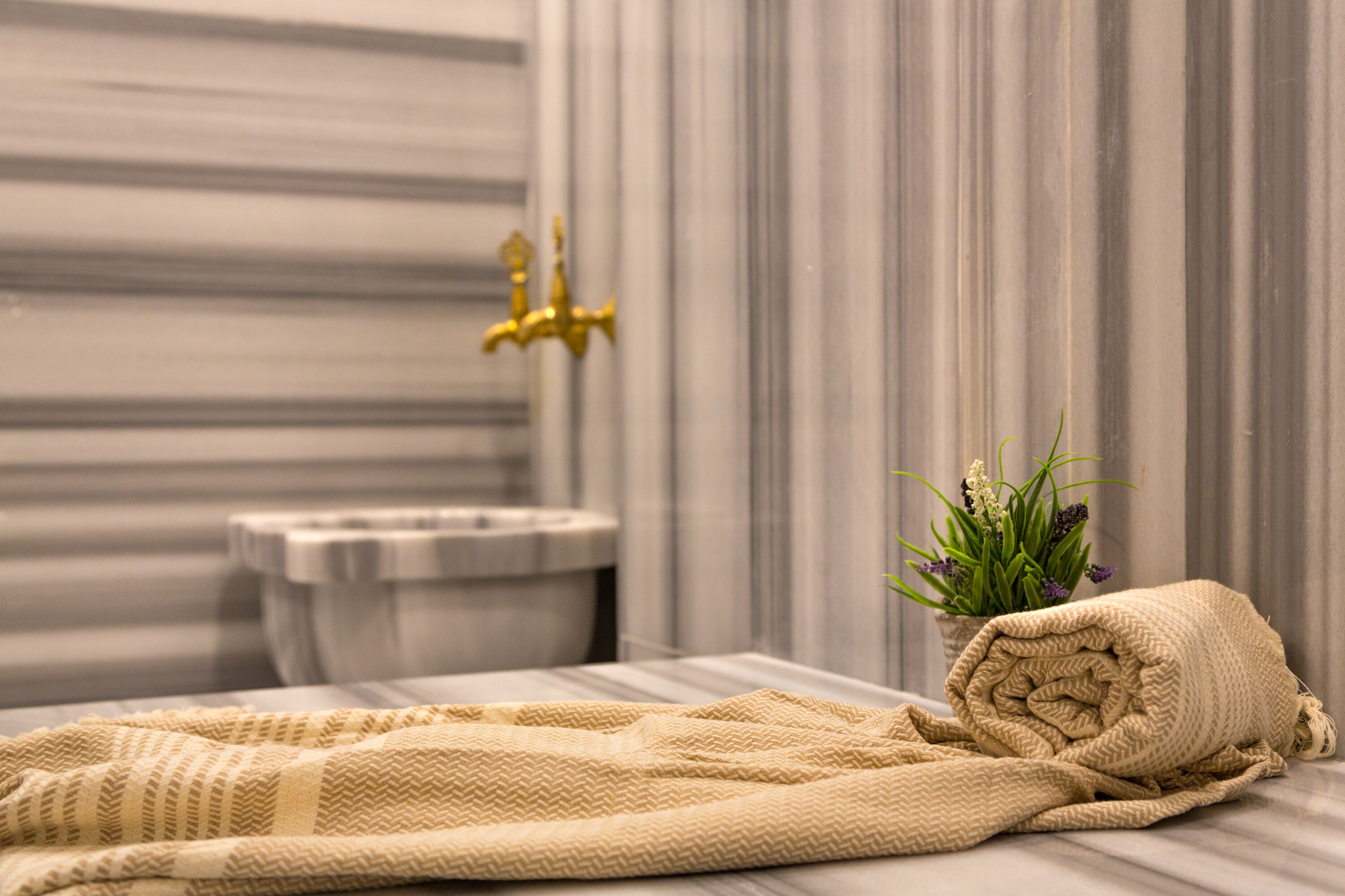 Une salle de bain avec un lavabo en marbre, un robinet doré, une serviette beige et une plante en pot ornée de fleurs vertes et violettes.