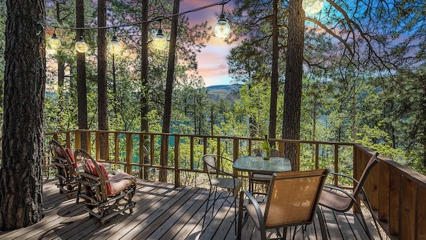 Une terrasse en bois, agrémentée de chaises et d’une table en osier, offre une vue sur une zone boisée et sur des collines lointaines.