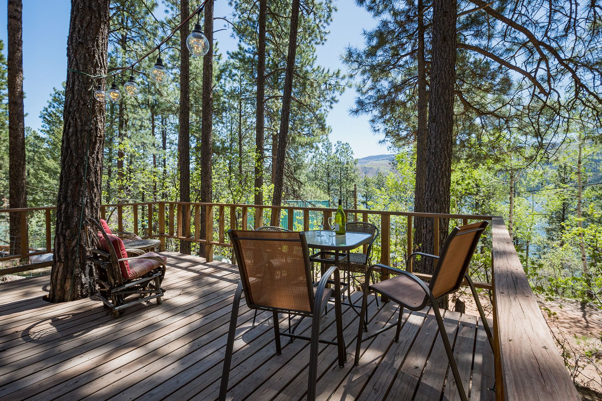 A wooden deck with a table and chairs, surrounded by tall pine trees.