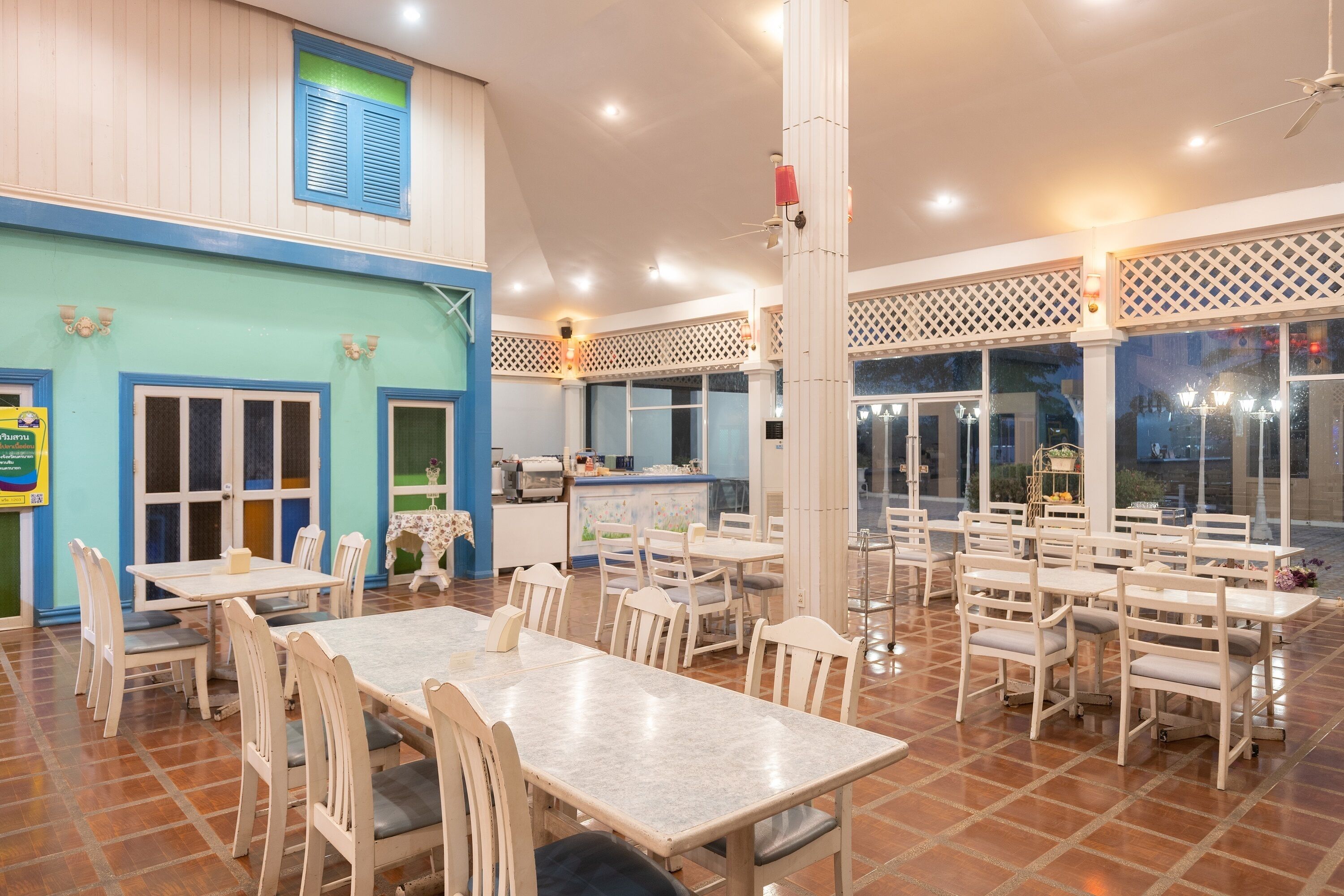 A spacious indoor dining area with multiple tables and chairs, a kitchen area in the background, and large windows allowing natural light.
