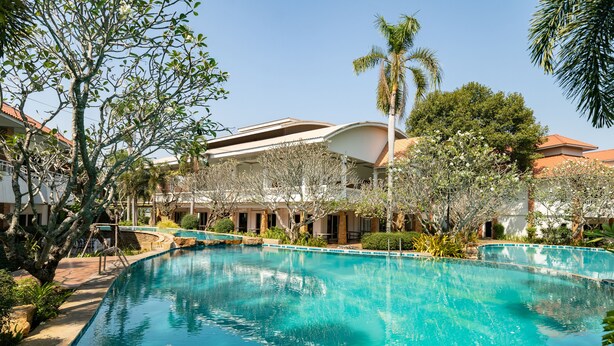 A swimming pool surrounded by buildings with a curved roof, palm trees, and a clear blue sky. - Thara Cholapruek Resort (Ban Na)