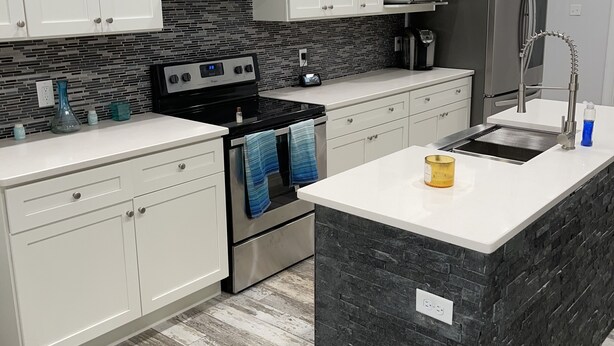 Une cuisine moderne avec des meubles blancs, un îlot central, des appareils en acier inoxydable et un dosseret de carrelage disposé en motif chevron.
