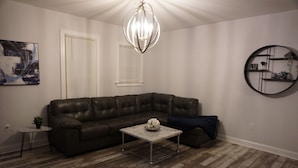 Un salon comprenant un canapé d’angle en cuir, une petite table basse ronde, un lustre, une étagère murale ronde et un tableau accroché au mur.