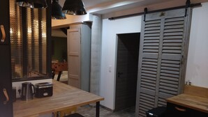 A kitchen with a wooden table, two stools, a coffee maker, and a toaster. There is a sliding door with horizontal slats and a black trash bin. - Charming house with swimming pool (Saint-Chamond)