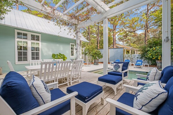 A covered outdoor seating area with white chairs and blue cushions, a pool, and a house with a green exterior.